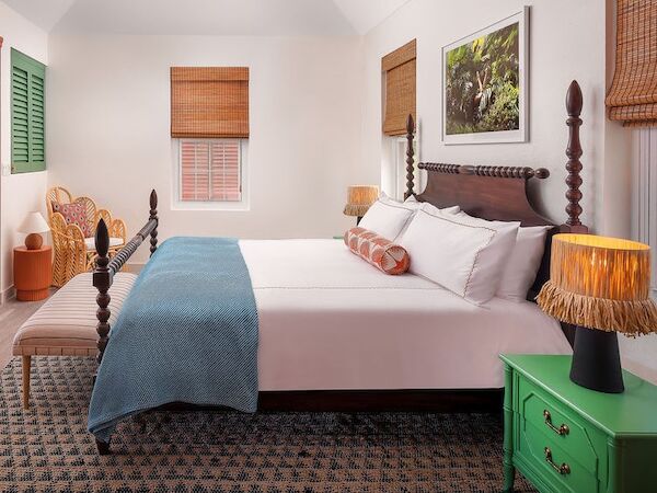 A cozy, well-lit hotel room with a large bed, teal throw, green nightstand, wicker blinds, and warm lamps—comfortable and inviting.