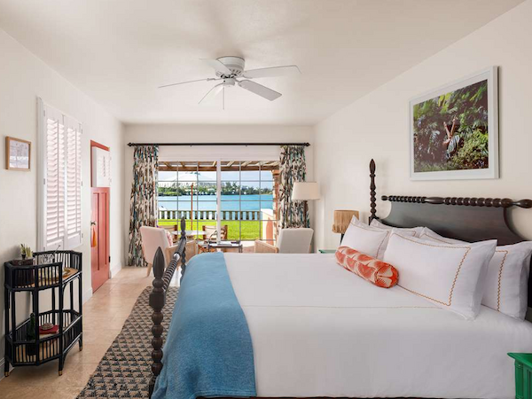 A bright hotel room with a king bed, white linens, blue throw, ceiling fan, desk, seating area, and a view of a patio and water beyond.