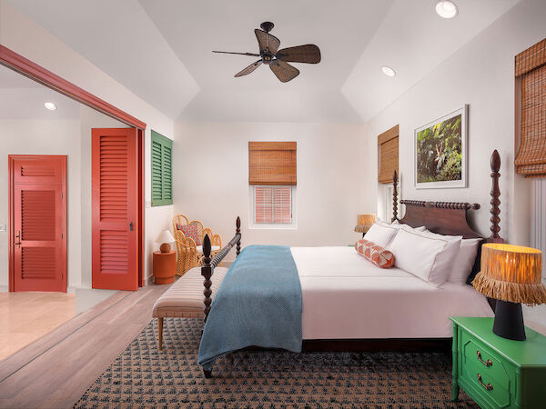 A cozy bedroom features a double bed with a wooden frame, colorful doors, wicker furniture, a green nightstand, and wicker blinds on the windows.