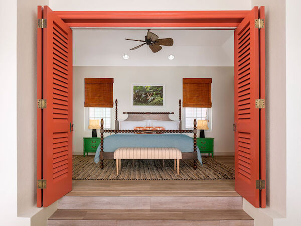 A cozy bedroom with a large bed, orange shutters, and green accents. The room is well-lit and features wooden blinds, a ceiling fan, and side tables.