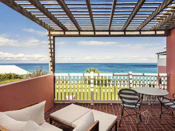 A patio with wooden furniture overlooks a grassy area, picket fence, and expansive ocean view. The sky is partly cloudy, providing a serene setting.