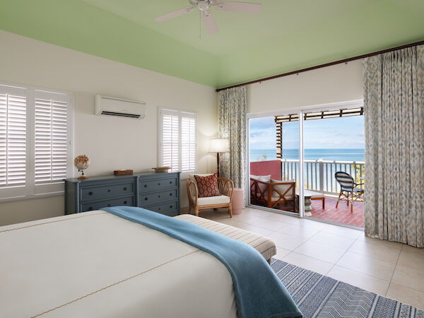A cozy bedroom featuring a large bed, a chest of drawers, a chair, a lamp, and an open balcony with a view of the ocean.