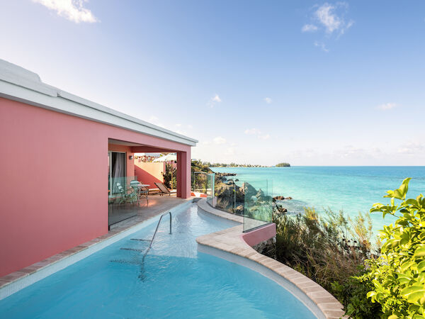 A picturesque oceanfront villa with a private pool and pink exterior, overlooking a clear blue sea under a sky with scattered clouds.
