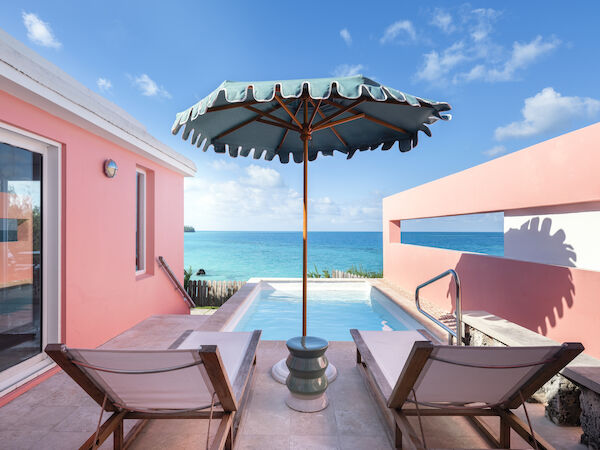 Image shows two lounge chairs under a large umbrella next to a pool, with an ocean view, flanked by pink walls under a clear blue sky.