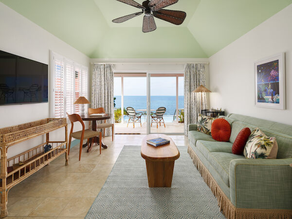 A cozy living room with green accents features a sofa, coffee table, desk, TV, and opens to an ocean-view balcony with chairs and a table.