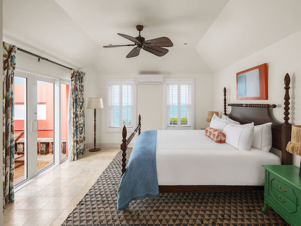 A spacious bedroom features a large bed, a ceiling fan, a green nightstand, and glass doors leading to a balcony with an ocean view.