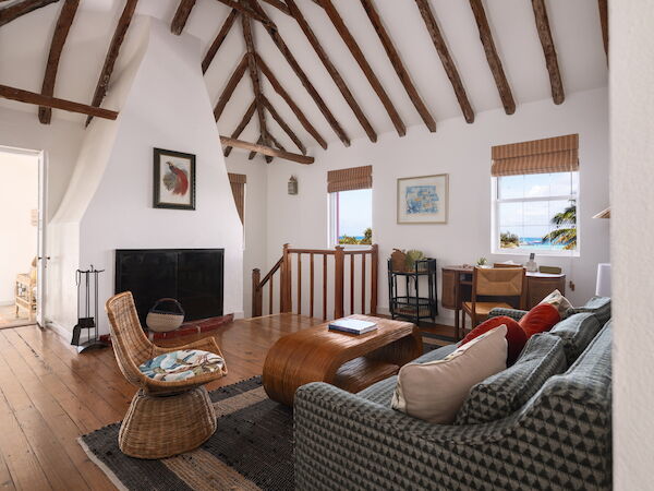 A cozy living room with rustic beams, a fireplace, a sofa, a wicker chair, a coffee table, and artwork, overlooks a scenic view through two windows.