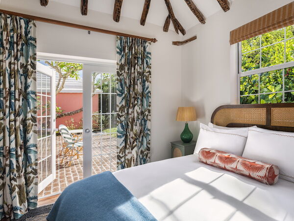 A cozy bedroom with exposed beams, a comfy bed, patterned curtains, a lamp, and French doors opening to a sunny patio with outdoor seating, ends the sentence.