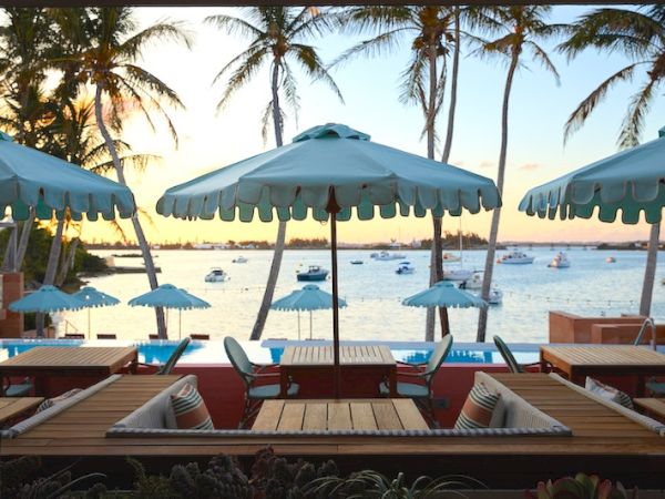 The image shows a scenic view of an outdoor seating area with umbrellas by the water, palm trees, and boats in the background at sunset.