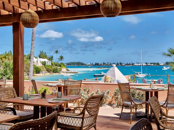 An outdoor dining area with wooden tables and wicker chairs overlooks a scenic beach with blue water, sailboats, and a clear sky.