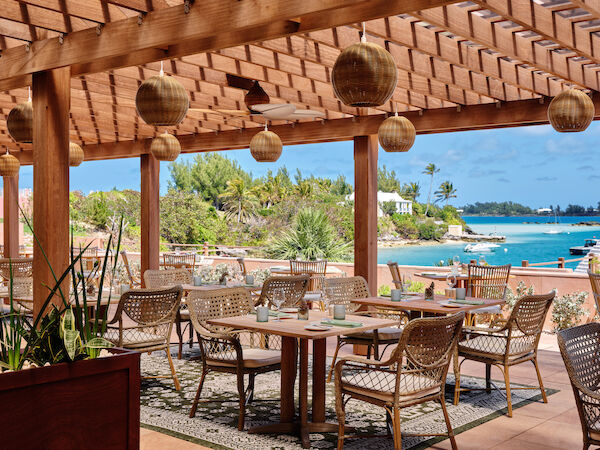 An outdoor restaurant or cafe with wicker chairs and wooden tables under a pergola, overlooking a serene waterfront with lush greenery.