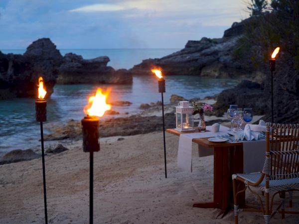 A romantic beachside dinner setup with torches, a table for two, and soft-lit lanterns, set against a backdrop of rocky shore and serene water.
