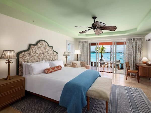 The image shows a spacious bedroom with a large bed, tropical-style decor, and a balcony with outdoor seating that overlooks the ocean.
