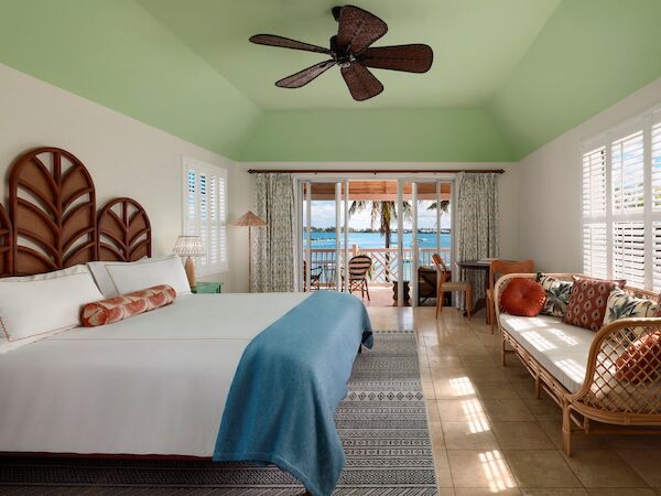 This image shows a bright and airy bedroom with a large bed, a ceiling fan, a couch, and a window with a view of the ocean and palm trees outside.