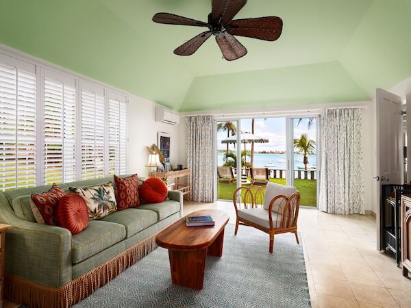 A modern tropical living room with a ceiling fan, green sofa, wooden furniture, and large windows opening to a patio with a beach view ending the sentence.