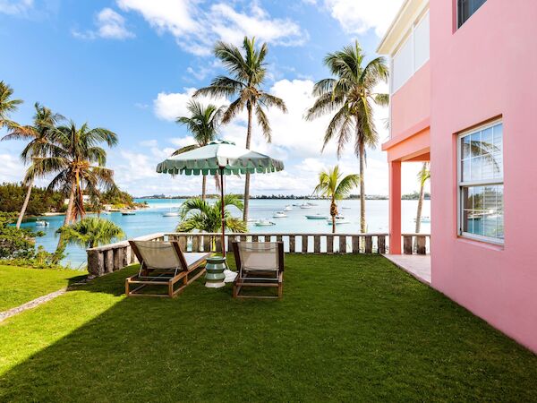 A seaside scene with two lounge chairs under a striped umbrella, surrounded by palm trees, a pink building, and a view of boats and the ocean.
