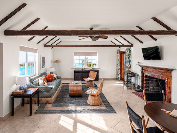 A cozy living room with exposed beams, a fireplace, a TV, and rattan furniture. There are multiple windows bringing in natural light.