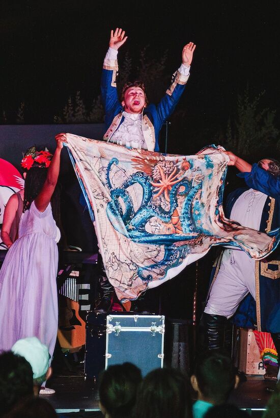 A theatrical performance with actors in costumes, one holding a colorful cloth with an octopus design, and another raised hands on stage.