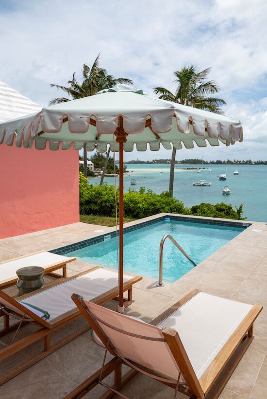 A poolside setup with lounge chairs and an umbrella overlooks a calm ocean view with boats, surrounded by lush greenery and palm trees.