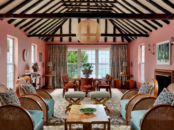 A cozy living room with a pink color scheme, featuring wooden beams, wicker furniture, a large central table, and windows with lush outside views.