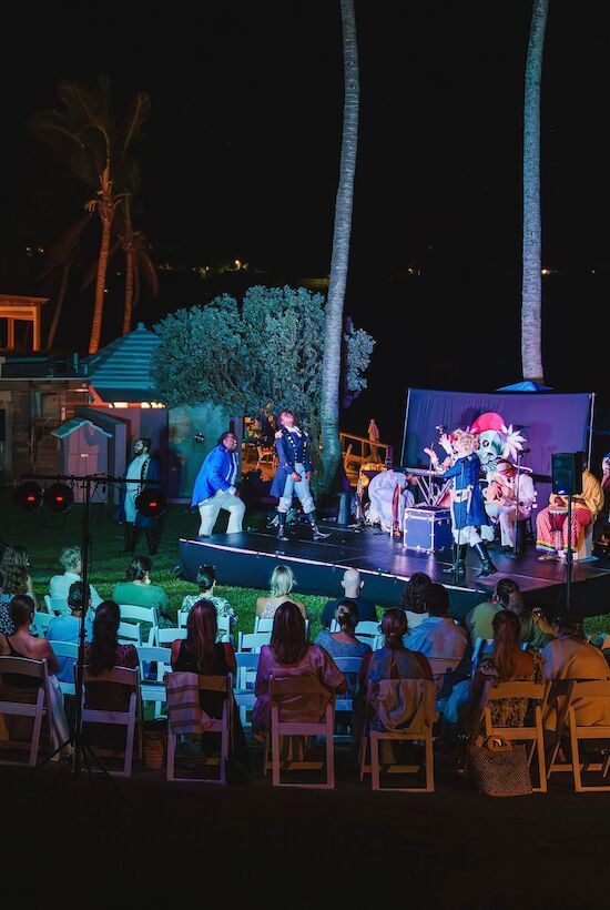 An outdoor stage performance at night with an audience seated in front, surrounded by palm trees and illuminated by colorful lights.