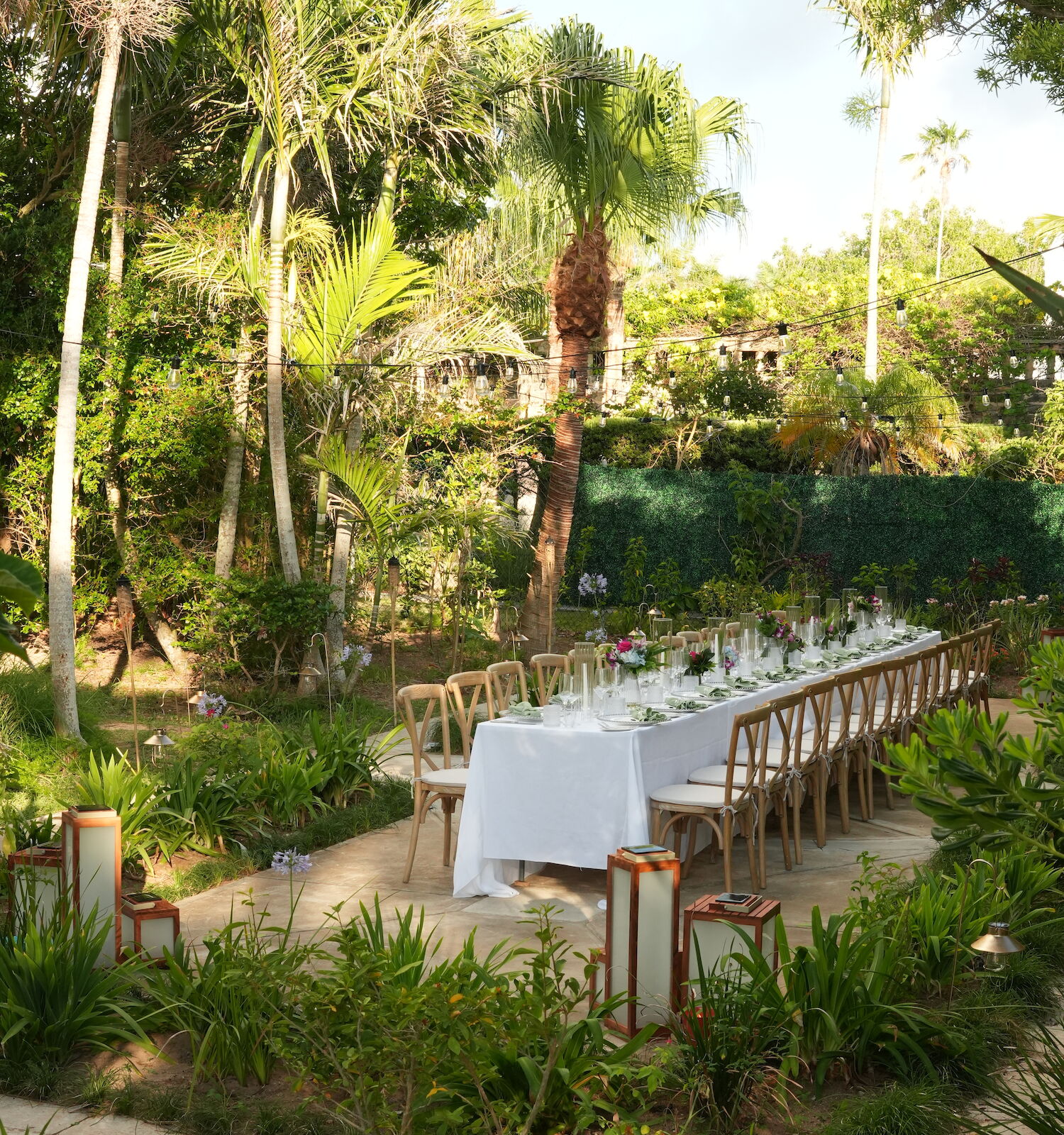 A long outdoor dining table with white tablecloths is set in a lush garden surrounded by palm trees and greenery.