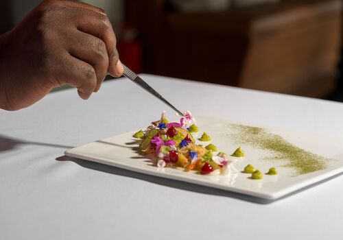 A hand uses tweezers to arrange a colorful, artistic dish on a white rectangular plate, showcasing a fine dining presentation.