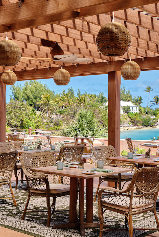 An outdoor dining area by the water, featuring wooden furniture, woven pendant lights, and a view of lush greenery and a clear sky.