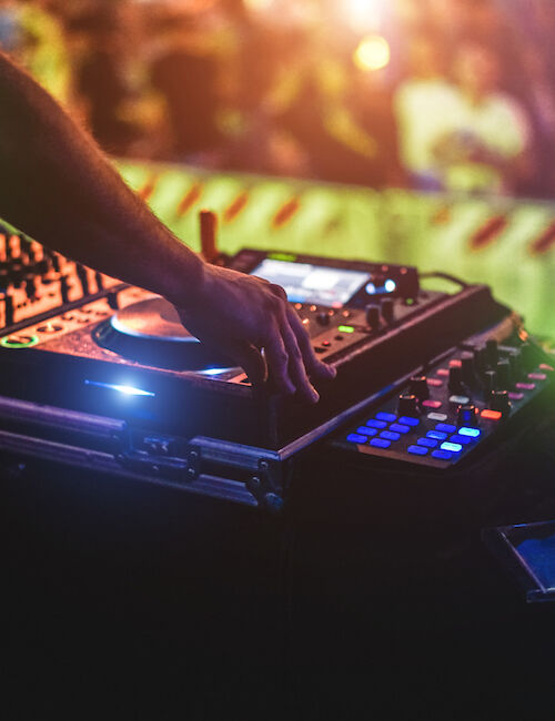 A DJ is mixing music using turntables and a laptop in a lively, colorful setting, with a blurred crowd in the background.