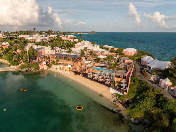 Aerial view of a coastal resort with a sandy beach, pool, loungers, umbrellas, and nearby houses by the ocean.