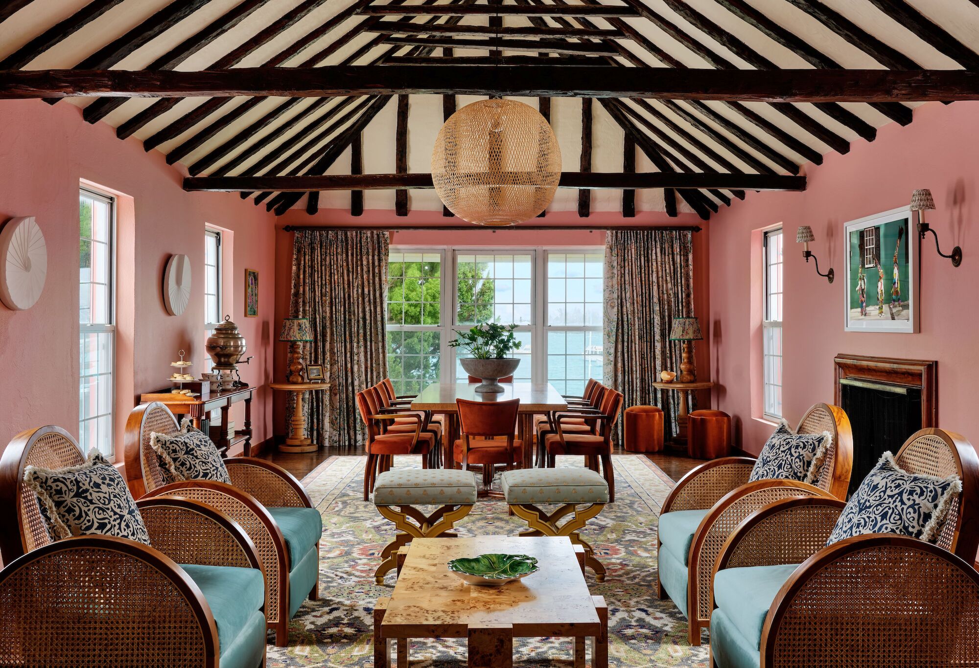 A cozy living room with pink walls, exposed beams, and eclectic decor. Featuring wooden furniture, cushioned seating, and large windows.