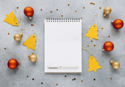 A spiral notebook and pen are surrounded by gold and red Christmas ornaments and tree-shaped tags on a gray surface, with scattered confetti.