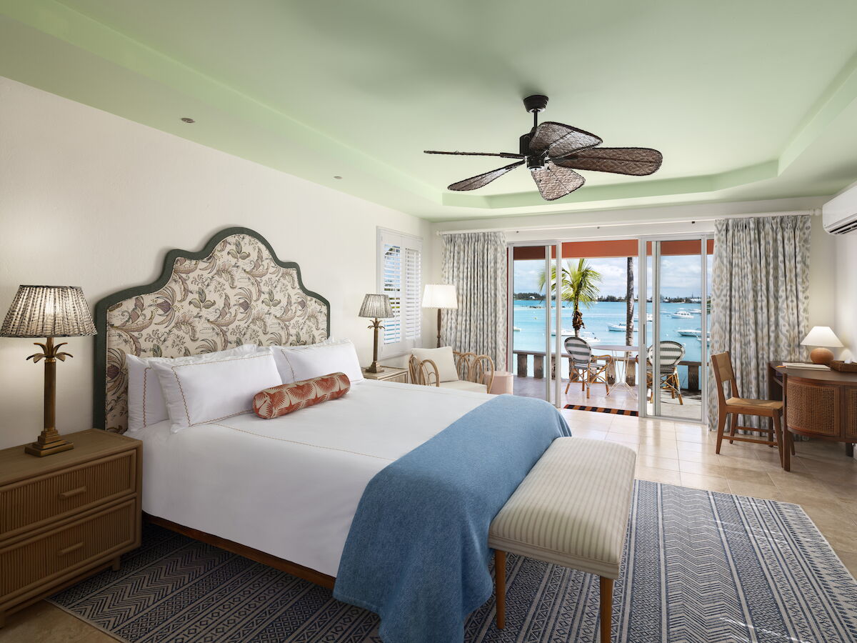 A bright bedroom with a large bed, ceiling fan, ocean view, and outdoor seating on a balcony, creating a relaxing atmosphere.