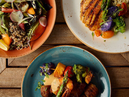 The image shows three plates of food with grilled items, vegetables, and quinoa salad on a wooden table.