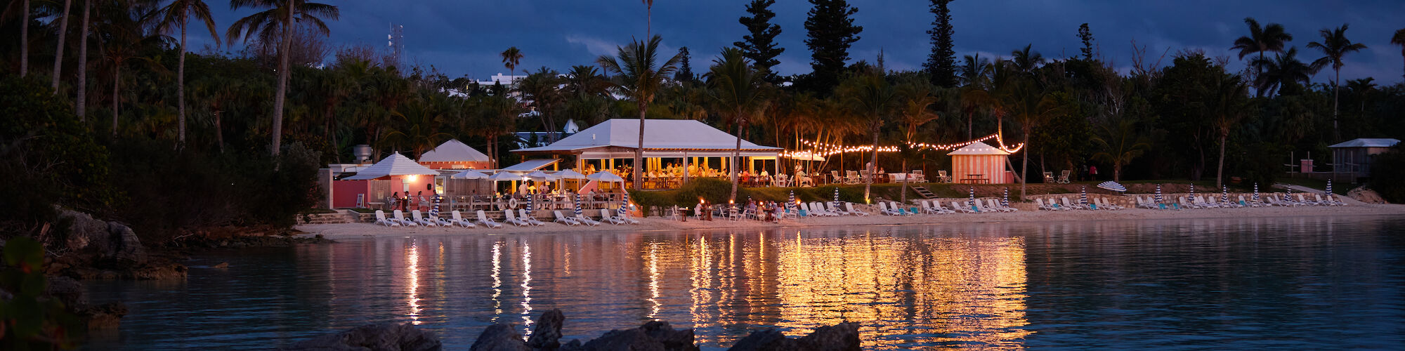 A beach resort at dusk with illuminated buildings, palm trees, and a calm sea reflecting the lights, creating a tranquil atmosphere.