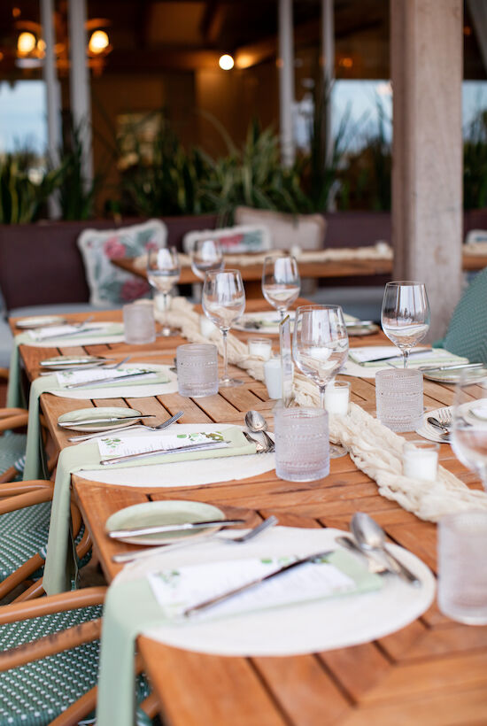 A wooden table is set for a meal with white place settings, glasses, utensils, and light green napkins in a cozy outdoor setting.