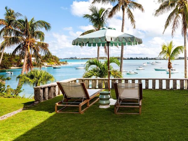Two lounge chairs and an umbrella sit on a grassy area overlooking a tropical beach with palm trees and boats on the water.
