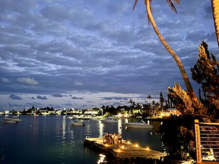 Scenic dusk view of a waterfront with boats, palm trees, and illuminated docks under a cloudy sky.