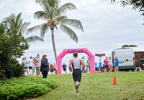 A person is approaching the pink "FINISH" arch at an outdoor event, with spectators and palm trees in the background.