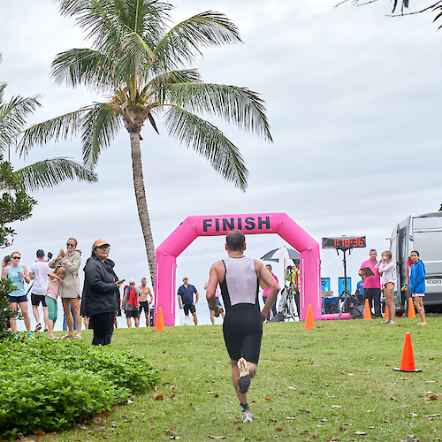 A person is approaching the pink "FINISH" arch at an outdoor event, with spectators and palm trees in the background.