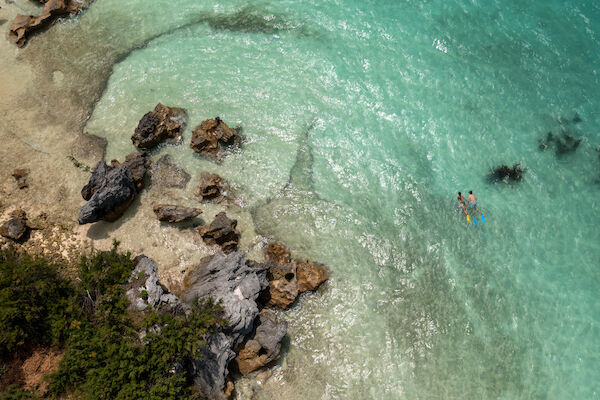 An aerial view of clear turquoise water with rocks and two people swimming near the shore.