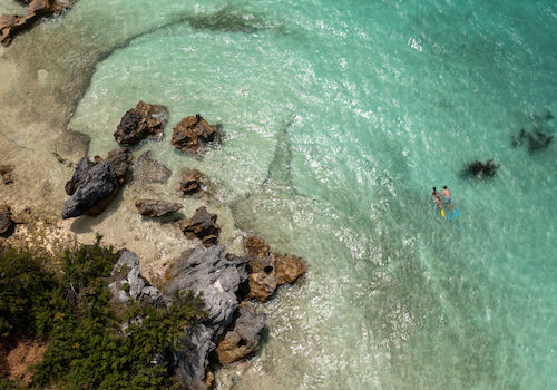 An aerial view of clear turquoise water, rocks, and two people swimming near the shore, surrounded by a natural coastal landscape.