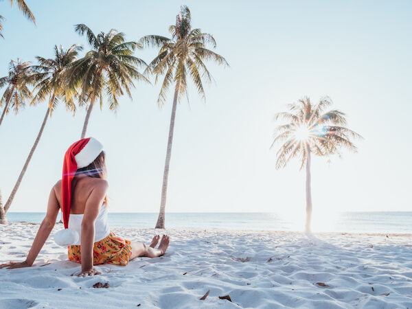 A person in a Santa hat sits on a sunny beach with palm trees and looks out at the ocean.