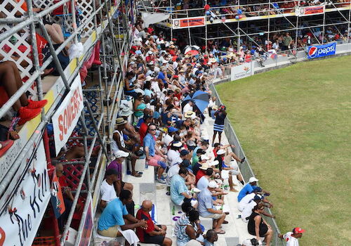 A large crowd is seated in stadium stands, watching an event on a grassy field, with structures and palm trees in the background.
