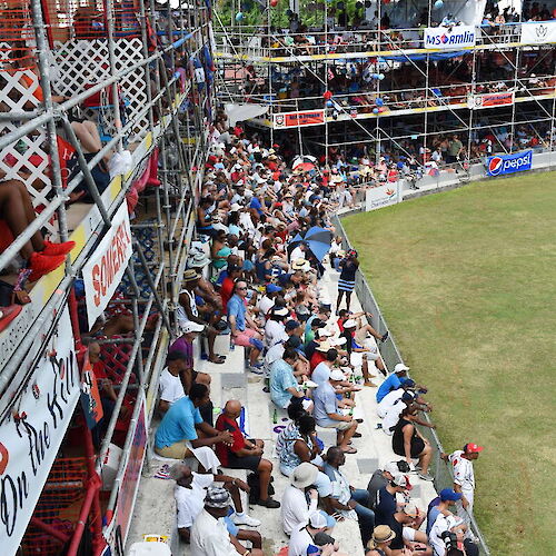 A large crowd is seated in stadium stands, watching an event on a grassy field, with structures and palm trees in the background.