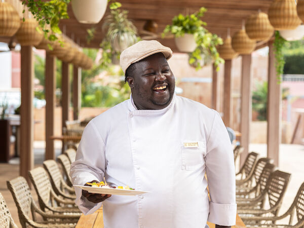 A person in chef attire holds a dish in an outdoor dining area with wooden tables and plants.