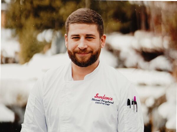 A person in a white chef's coat with pens in the pocket, standing outdoors in a snowy setting, smiling at the camera.