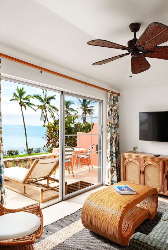 A tropical-themed living room with a sea view, wicker furniture, patterned curtains, ceiling fan, and deck with lounge chairs outside.