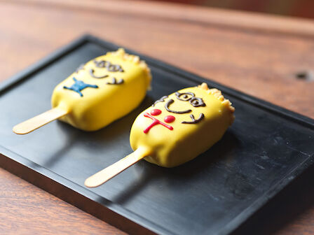 Two ice cream bars on sticks, coated in yellow with cartoon character designs, placed on a black tray.