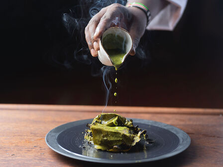 A person pours a green sauce over a plated dish, with steam rising from the food, on a wooden table against a dark background.